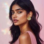 A woman wearing gold-tone metal earrings with a double-layered circular design, resembling the sun's rays, set against a pink background with bokeh lights.