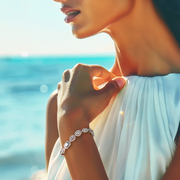A close-up image of a woman's wrist wearing a silver bracelet with a cascading pattern of zirconia stones, set against a beach backdrop.